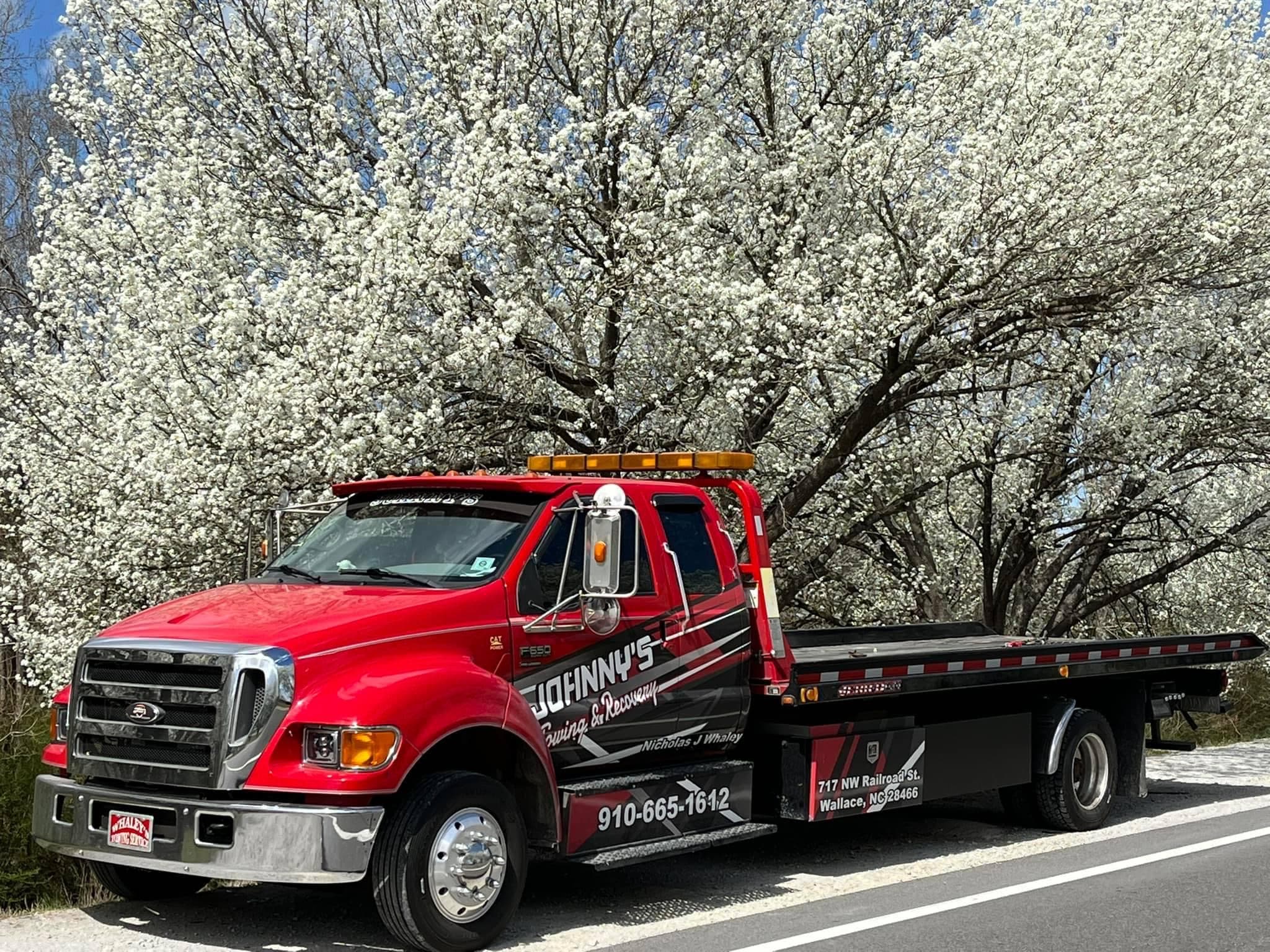 Johnny's Towing & Recovery red flatbed truck parked in front of flowering trees in Wallace, NC